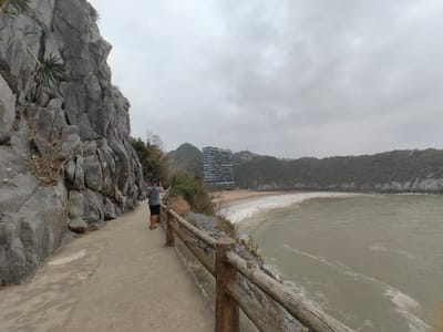 A wide-angle shot depicts a coastal scene on an overcast day in Hải Phòng, Vietnam. A man stands on a concrete walkway, facing the ocean and holding a mobile device to capture a photograph. The walkway is bordered by a weathered wooden railing and a rocky cliff face on the left side. The water of the bay is murky, with visible wave action. In the background, a multi-story building with a unique, colorful facade stands on a hilltop overlooking a sandy beach. Additional forested hills and a cloudy sky complete the composition.