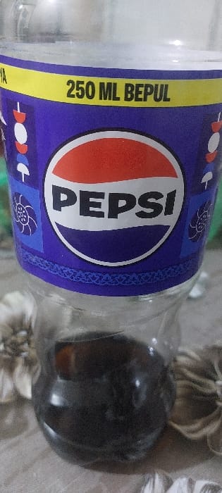 A clear plastic beverage bottle is depicted upright, partially filled with a dark brown liquid occupying approximately the bottom third of its volume. The bottle features a purple label with a prominent yellow horizontal band across the top, displaying the text "250 ML BEPUL" in black capital letters. Below this band, the classic circular Pepsi logo, consisting of red, white, and blue sections with "PEPSI" in white capital letters, is centrally positioned. Decorative patterns, including a small white mushroom-like icon and a stylized blue floral motif, are visible on the purple label. A subtle patterned border is present along the lower edge of the purple section of the label. The bottle rests on a light-colored, textured surface, possibly fabric. Blurred, light-brown objects, resembling artificial flowers or decorative elements, are visible in the background on both sides of the bottle. No individuals or specific actions are depicted. The scene is situated in Nazarbek, Uzbekistan, a detail informed by the provided context and the Uzbek word "BEPUL" visible on the label.