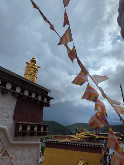 The image captures a view of the Songzanlin Monastery in Shangri-La, China, under a dramatic, cloudy sky. In the foreground, a section of the monastery's white and red plastered wall is visible, featuring decorative white circles and a dark red upper facade. Prayer flags, in alternating stripes of purple, yellow, orange, and white, are strung across the sky, some appearing weathered and torn.

Dominating the left side of the frame is the golden stupa atop one of the monastery's roofs, intricately detailed and reflecting the ambient light. Below this, another building features a similar architectural style with dark trim and white decorative elements.

In the background, the extensive golden roofs of the Songzanlin Monastery complex spread across the landscape, with several tiered structures topped with golden spires. Lush green hills rise behind the monastery, disappearing into the misty, overcast sky. A few people are visible on the monastery grounds in the distance, appearing as small figures against the grand architecture. The overall scene suggests a serene, yet powerful, spiritual site set against a dynamic natural backdrop.