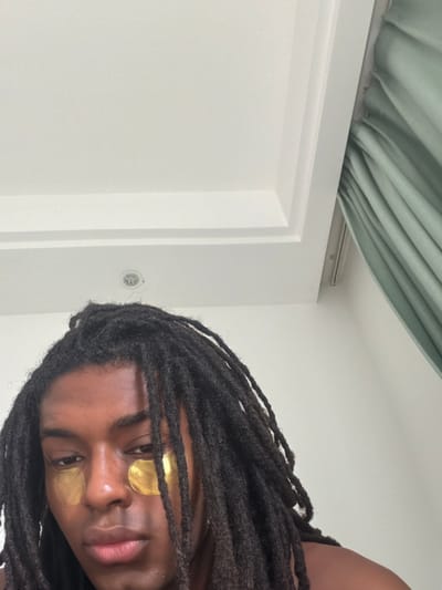A medium shot depicts a person with locs and gold eye masks in a room with white walls and a pale green curtain. The location is unknown. The person is facing the camera, tilted to the right. The background includes a closed white door with a round, metallic fixture on the wall nearby.