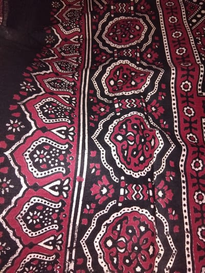 This is a close-up, top-down, eye-level, full-frame shot of a piece of fabric with a traditional block-printed design. The fabric is predominantly black with intricate patterns in deep red and white. The design features repeating geometric and floral motifs arranged in vertical bands. One band consists of a series of stylized floral shapes outlined in white and filled with red and black. Adjacent to this is a wider band with larger, elaborate circular motifs, also outlined in white and filled with red and black, interspersed with smaller white and red floral elements. Another vertical band features a simpler pattern of alternating red and white shapes with black outlines. The fabric appears to be slightly creased, suggesting it is a textile likely used for clothing, upholstery, or as a decorative textile. The image does not depict any people or identifiable landmarks, and therefore no specific location within Haveli Jhangranwali, Pakistan can be determined.