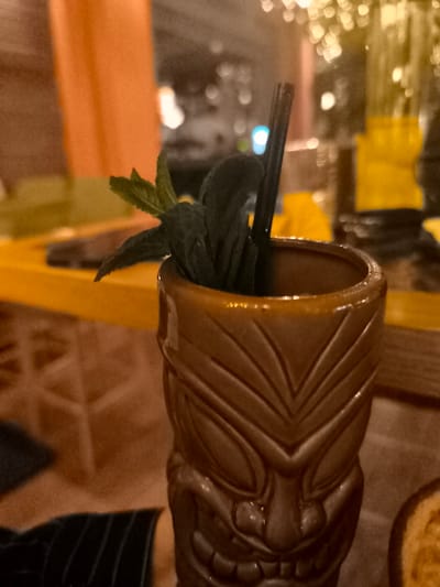 An indoor, close-up shot features a tiki mug cocktail with mint garnishes and a black straw, set against a blurred background of warm, artificial lighting. The focal point is a brown, textured ceramic tiki mug, designed with an intricate, stylized face motif visible on its lower half. Fresh green mint leaves are clustered at the top of the cocktail, protruding from the mug, alongside a slender black drinking straw.

The background is significantly out of focus, suggesting a shallow depth of field. It shows glowing bokeh lights, predominantly in warm yellow and orange hues, with a singular brighter blue light appearing on the left side of the straw. Hints of a yellow surface, possibly a bar top or table, are visible behind the mug. To the far left, an orange-colored element, possibly a wall or curtain, is present. In the lower left corner, a person's arm, dressed in a dark fabric with subtle stripes, is partially visible, indicating human presence at the table.

The overall lighting is dim and atmospheric, suggesting the photograph was taken in the evening or at night in a bar or restaurant setting, perhaps in Palaio Faliro, Greece. There is no visible text in the image.