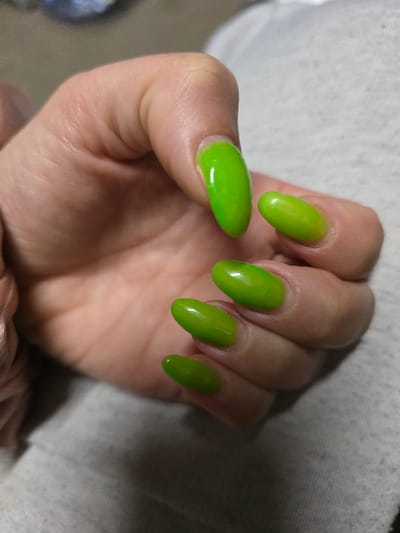 A close-up image depicts a human left hand, positioned over a light grey, textured surface. Five fingers are visible, each terminating in an oval-shaped fingernail coated with a glossy, vibrant neon-green nail polish. Minor nail regrowth is visible at the cuticle line of several nails. The skin of the hand is fair, exhibiting natural creases and folds, particularly around the knuckles and palm. The hand is in a relaxed, slightly cupped posture. The background consists primarily of the light grey surface, with a blurred, lighter area visible in the upper left quadrant. The scene is located in Tetovo, Bulgaria.