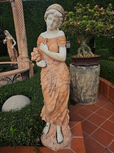 This image depicts an outdoor garden scene in Na Chom Thian, Thailand. The foreground is dominated by a life-sized marble statue of a woman in a flowing, orange-toned gown, holding a bouquet of flowers. The statue stands on a stone base, surrounded by lush green bushes and a large, smooth, gray boulder. To the left, partially visible, is another statue of a woman, also in an orange gown, positioned near a decorative stone pillar and a bench. In the background, a bonsai tree with pink flowers sits on a textured stone pedestal, set against a backdrop of meticulously trimmed green hedges. The ground is paved with reddish-brown tiles arranged in a diamond pattern. The lighting suggests it is daytime, with soft shadows cast on the ground.