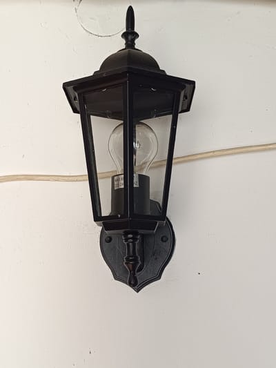 A black, vintage-style wall-mounted lantern is fixed to a textured white wall. The lantern has a pointed finial at the top, a hexagonal roof, and glass panes that reveal a clear, filament light bulb inside. A label on the bulb base reads "MAX 60W 220-240V 50HZ". A light-colored electrical wire runs horizontally across the wall behind the lantern. The wall has some minor imperfections and a circular mark above the lantern. The overall scene is static and appears to be an outdoor or semi-outdoor setting, potentially in Tebras, Latvia.