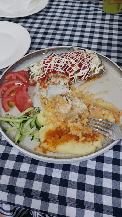 A plate of food is shown on a black and white checkered tablecloth. The plate contains mashed potatoes topped with a sauce, a meat dish with ketchup and mayonnaise drizzled on top, sliced tomatoes, and sliced cucumbers. A fork is stuck in the mashed potatoes. The setting appears to be a casual meal.