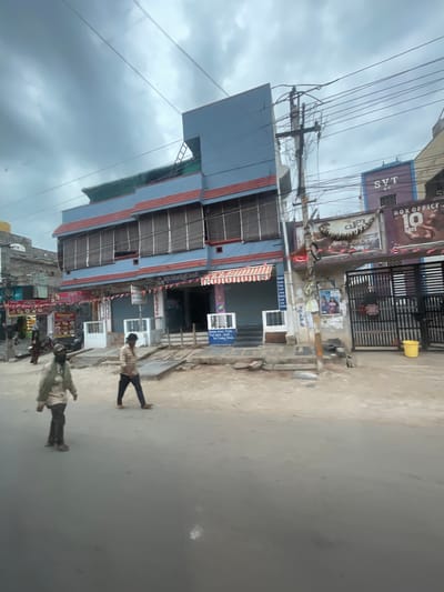 In Bethamcherla, India, the scene captures a street view with a multi-story building and its surroundings under a cloudy sky. The building, painted in blue and red, features multiple windows covered by blinds, suggesting commercial establishments on the lower floors. A sign with "SVT A/C" and "BOX OFFICE 10 DAYS" is visible on the right side of the building, indicating a cinema or entertainment venue. A metal gate is also present near this entrance.
In front of the building, two men are walking on the dusty street. One man, in the foreground on the left, wears a light-colored, possibly greenish, jacket and dark trousers. The other man, further to the right and slightly behind, is dressed in a checkered shirt and dark trousers. The street itself is unpaved and appears to be under construction or in a state of disrepair, with debris and wooden planks scattered about. A bright yellow bucket is placed near the gate.
The overall environment suggests a busy street in a developing urban area, with overhead power lines crisscrossing the sky, a common feature in many Indian cities. The weather appears overcast, contributing to a muted color palette of the scene.
