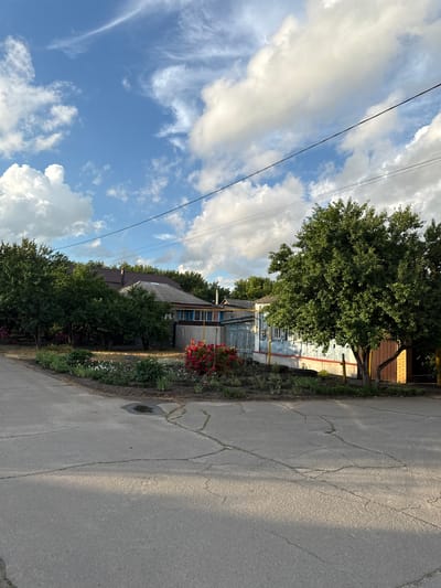 The image depicts a street scene in the city of Rossosh, Russia, featuring a residential area with houses, trees, and a garden. The foreground shows a cracked asphalt road with a shadow cast across it, indicating sunlight. To the left, a flowerbed with red roses and other plants is visible, bordering the road. Behind the flowerbed are several trees with lush green foliage.
In the mid-ground, a light blue house with a brown roof and white trim is partially visible, with a wooden fence in front of it. A large, mature tree with dense green leaves stands prominently to the right of the house. Power lines are strung across the sky.
The background consists of more trees and the sky, which is a vibrant blue with scattered white cumulus clouds. The overall scene suggests a sunny day in a rural or suburban environment.