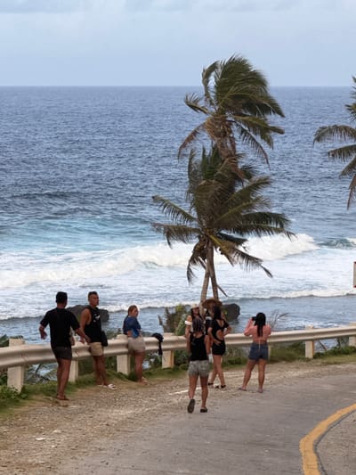 The image shows a group of people standing on a road next to a white guardrail, with the ocean visible in the background. Two palm trees are in the foreground between the road and the ocean. Six people are lined up on the side of the road, and two people are walking further up the road away from the camera. The road appears to have a yellow painted line. The scene is likely near the coast in Burgos, Philippines, given the vegetation and ocean view.