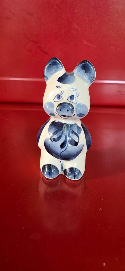 A small, white ceramic figurine of a pig with blue floral patterns is placed on a red, reflective surface. The pig has a rounded body, short legs, and large ears. Its face is adorned with blue painted eyes, eyebrows, cheeks, and a snout. A blue floral design is painted on its chest, resembling a bow. The background is a solid red surface with a slightly textured appearance. The lighting is even, casting a subtle reflection of the figurine onto the surface below. The scene is indoors, likely on a table or shelf. There are no people visible in the image.