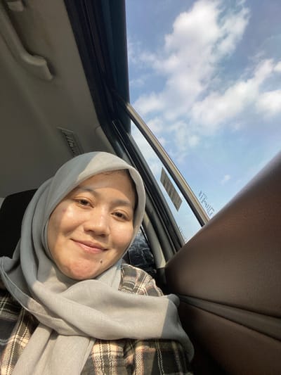 A woman wearing a light gray hijab and a plaid shirt is seated in the passenger seat of a car, looking directly at the camera. The car's interior is visible, with the ceiling and a portion of the passenger door. Through the open window, a bright blue sky with scattered white clouds is visible. The woman has a slight smile and her face is illuminated by sunlight. The image appears to be taken during the day. The location is Kertak Hanyar I, Indonesia.
