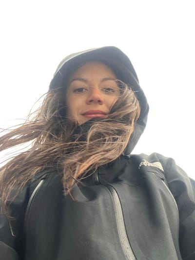 A woman in a black hooded jacket looks into the camera, her head tilted slightly. She has long, wet brown hair. The background is a bright, indistinct white. The jacket is zipped up, and the hood is pulled up around her head, framing her face. Part of the jacket's logo is visible on the right shoulder.