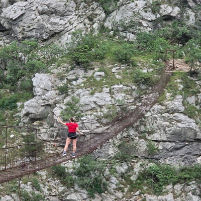 The image depicts a woman in a red shirt and dark shorts carefully traversing a rusty, wire-mesh bridge attached to a steep, rocky cliff face covered with sparse vegetation. The location is in the city of Unknown, Montenegro. The bridge appears to be suspended high above a likely canyon or steep ravine. The woman is holding onto the side of the bridge, suggesting it may be unstable or precarious. The environment consists of rugged terrain with large rocks and some greenery clinging to the stone.
