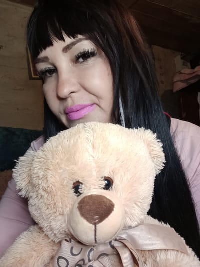 A selfie, taken from a high angle, shows a person lying down with a plush teddy bear. The person has long, dark hair, bangs, and is wearing a light pink shirt. Their face is partially obscured by the teddy bear, but their eyes are visible, with dramatic eyelashes and dark eyebrows. They are wearing bright pink lipstick. The teddy bear is light brown and fluffy, with a ribbon tied around its neck that has a pattern of grey circles and lines. The background is out of focus, but appears to be indoors with a textured wall and some furniture visible. The lighting suggests it could be daytime. The overall mood seems casual and playful. There is no visible text in the image. The location context is Sterlibashevo, Russia.