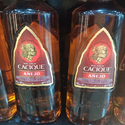 The image shows two bottles of "Ron Añejo Cacique" rum, a product of Venezuela, displayed side-by-side. The bottles are made of amber glass and have a distinctive label with a depiction of an indigenous person's profile, the brand name "Cacique," and "Añejo." Text on the labels indicates that the rum is elaborated by Destilerías Unidas, S.A., is registered under number L-69, and is made in the Bolivarian Republic of Venezuela. The alcoholic content is 40% G.L. and the net content is 0.75 L. The bottles are positioned in what appears to be a retail setting, possibly a store shelf, with reflections of lights visible on the glass. The image is cropped to focus on the labels and upper portions of the bottles. There are no people visible in the image. Based on the product's origin and the typical distribution of Venezuelan rum, it is plausible that this image was taken in Los Teques, Venezuela, a major city and the capital of the state of Miranda.