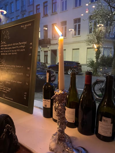 The image depicts an evening scene inside a restaurant looking out onto a street in Ixelles - Elsene, Belgium. In the foreground, on the left, a vertical black chalkboard menu with a green frame displays handwritten French menu items and prices, including "PAIN BEURRE 3.5" and restaurant name "@momebrussels". Visible entries include "ARTICHAUT, OSEILLE-CRESSON, POUTARGUE 16" and "MAGRET DE CANARD, ASPERGE BLANCHE ET VERTE, JUS CORSE, PDT 32". Centered, a lit yellow tapered candle with a bright flame stands in an improvised holder fashioned from crumpled aluminum foil, with white wax drips accumulated at its base. To the right of the candle, five wine bottles are arranged on a white surface, mostly dark green or black, with one labeled "BOAGNA" and another "BOURGOGNE BLANC 1750 ML". A black quilted handbag is partially visible on the lower left of the white window ledge.

Through the large window, the background reveals a street scene under artificial lighting. Across the street, multi-story European-style buildings with several lit windows are visible. Parked vehicles, including a dark blue SUV, line the street. A tree with sparse white blossoms is visible on the right, and a bicycle is partially obscured on the far right behind the wine bottles. Reflections of crescent-shaped interior lights are visible on the window glass, superimposed over the exterior view. The establishment is Môme Brussels, located at Rue de l'Aqueduc 13, 1050 Ixelles - Elsene, Belgium.