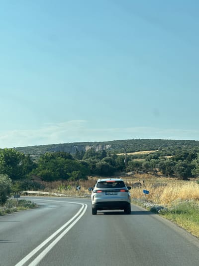 A silver SUV is driving on a paved road with double white lines in the city of Nea Triglia, Greece. The SUV is viewed from behind, with its taillights illuminated. The road curves to the left, with a guardrail on the left side of the image. The surrounding environment is rural, with dry grass, olive trees, and hills in the background. A road sign indicating "49" is visible on the right side of the road. The sky is clear and blue.