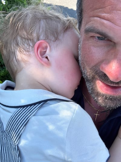 An intimate close-up captures a man and a young child resting together outdoors in Bredene, Belgium, under bright daylight. The image is taken from an overhead perspective, with the subjects positioned diagonally.
The man, visible in the upper portion of the frame, has light skin, a greying beard with dark stubble, and a partially visible blue-grey eye. He wears a dark blue top, and a prominent gold chain with a small gold cross pendant is visible around his neck. His face is positioned closely to the child's.
The child, appearing very young, rests their head near the man. Only a portion of their light-skinned face, ear, and light blonde or light brown hair are visible. Their eyes are closed, suggesting they are asleep or peacefully resting. The child's cheeks have a slight rosy flush. They are dressed in a white polo shirt, featuring a distinctive navy blue and white striped pattern on the inner collar or placket.
The bright sunshine illuminates the scene, casting soft shadows and highlighting details like the texture of the man's beard and the child's hair. Glimpses of green foliage and a small white flower in the bottom left corner indicate a grassy outdoor setting. The overall mood conveyed is one of tenderness, affection, and peaceful rest. There is no visible text in the image.