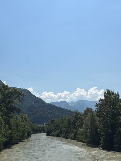 The image captures a wide, muddy river flowing through a valley with forested hillsides. The sky is a clear blue with a few fluffy white clouds. In the distance, a range of majestic mountains is visible. The vegetation along the riverbanks is dense and green, with a variety of trees. The river itself appears to be flowing at a moderate pace, with some ripples and reflections on its surface. A town or village can be seen nestled among the trees on the far bank, with a few buildings visible. The overall impression is one of a serene and natural landscape, likely situated in a mountainous region with a substantial river. This scene is consistent with views of the Arve River in or near Cluses, France, a town located in the Alps.
