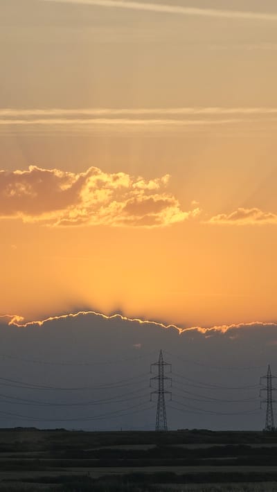 This is a vertical, low-angle photograph taken at sunset. The scene depicts a landscape with a dramatic sky and man-made structures. The left side of the image is dominated by a pale, warm-toned sky with faint wispy clouds or contrails. Moving towards the center and right, the sky transitions to a vibrant orange, illuminated by the setting sun. A large, textured cloud formation is visible, catching the light and appearing bright yellow and orange.

The right side of the image is darker, with a silhouette of power lines and two tall electrical pylons. These structures stretch across the frame, against the backdrop of the darkening sky and a strip of land at the very edge, which appears to be fields or agricultural land. The contrast between the brightly lit sky and the shadowed landscape is striking. The setting sun creates a glowing edge along the horizon where the clouds meet the darker sky. The overall mood is serene and slightly dramatic, enhanced by the warm, fading light and the imposing presence of the power lines. The time of day is clearly sunset, given the intense colors in the sky and the long shadows that would be cast. There is no visible text in the image. The location context provided is Muel, Spain.