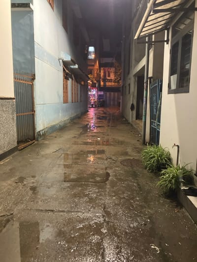 The image displays a narrow, wet concrete alleyway at night, located in Ngũ Hành Sơn Ward, Vietnam. The ground is uneven, featuring numerous puddles that reflect colorful artificial lights, predominantly pink, red, and orange, from the alley's distant end. On the left, a multi-story building painted light blue has multiple rectangular windows with wooden frames and a rusty, vertical-bar metal gate leading into an entryway. The lower section of this building's wall is finished with a grey, speckled material. The right side of the alley is bordered by a lighter-colored building, which features a sloped, corrugated awning supported by metal struts. Further along this side, a blue metal gate is visible. Two potted green plants are positioned near the right-hand building, adjacent to a wall-mounted water tap. A circular metal manhole cover is present on the wet concrete surface in the foreground. No individuals or vehicles are present in the scene.