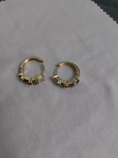A pair of gold hoop earrings with multi-colored gemstones are laid out on a textured light-colored fabric. The earrings are on a flat surface, not being worn. There are no people visible in the image, and no specific location information is provided within the image itself. Therefore, it is not possible to determine if this scene is located in Жарлыкөл, Kazakhstan.