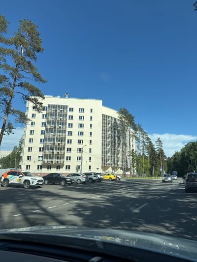 This image captures a street scene on a clear, sunny day in Pesochny, Russia. The perspective is from inside a vehicle, looking forward through the windshield.
In the left foreground, a tall pine tree with green needles and a brown trunk partially frames the scene. Behind it, a light-colored, multi-story building, likely a residential complex, dominates the left midground. This building features numerous rectangular windows with white frames and a prominent, multi-story glass-enclosed section on its facade. The rooftop has visible ventilation or communication equipment.
Adjacent to the building, a paved area serves as a parking lot, where several vehicles are parked. In the immediate left foreground, a white Toyota RAV4 SUV with orange and green "ОР" branding and partially visible Russian license plate (e.g., "У105ОМ 198") is parked. A black sedan, a silver sedan, and another white SUV are parked further along.
The central and right portions of the image are occupied by an asphalt road with visible white dashed lane markings. Strong, elongated shadows from unseen trees or structures are cast across the road and parking area. Traffic is present in the distance, with a silver SUV and a white sedan visible in the right lane, heading away from the camera. A blue truck is also visible further down the road. On the extreme right, a dark SUV is parked on the shoulder, facing away from the viewer.
A dense line of tall pine and deciduous trees borders the right side of the road, extending into the background. A single street light pole is visible between the building and the treeline. The sky above is a clear, deep blue with a few small, wispy white clouds visible in the upper right. No individuals are clearly discernible in the image.