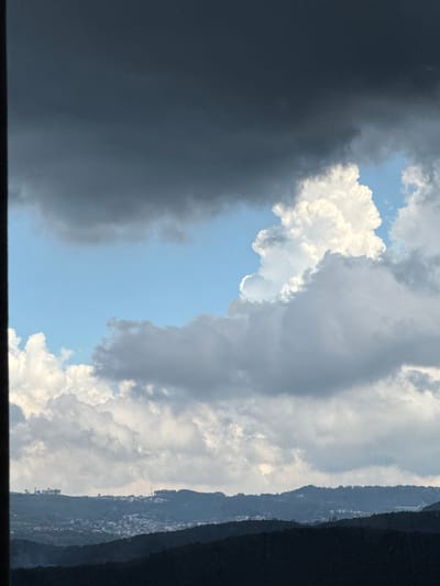 The image shows a cloudy sky over a hilly landscape with a town nestled in the valleys. The sky is filled with various types of clouds, ranging from dark, heavy storm clouds to bright, puffy white cumulus clouds. In the distance, rolling hills covered in dark green foliage form the horizon. A town with buildings of various sizes and colors is visible on the slopes of the hills, with some structures appearing to be houses and others possibly commercial buildings. The overall impression is of a serene, natural environment with a human settlement integrated into the landscape. The specific location in Đa Lộc, Vietnam, is not explicitly identifiable from the image alone, but the terrain and vegetation suggest a tropical or subtropical climate.