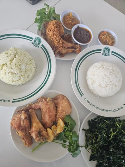 A top-down, slightly angled shot captures a meal spread out on a white, tiled surface. The meal consists of several dishes, primarily featuring fried chicken and rice, with accompanying condiments and vegetables.
In the upper center, a plate holds a generous portion of fried chicken, coated in crispy crumbs and appearing seasoned. Beside it, a small plate is laden with three small bowls of chili paste or sambal, varying in color from light orange to a deep reddish-brown. A sprig of fresh green leaves, possibly watercress, garnishes this plate.
To the left, a white plate with green trim showcases a mound of fluffy white rice, speckled with what appear to be herbs. The same branded plate with green trim is visible on the right, also holding a portion of white rice.
Below these, another white plate displays an assortment of fried chicken pieces, including wings and drumettes, alongside some fried garlic. A pile of dark green, wilted leafy vegetables is presented in a separate white dish on the lower right.
The branding on the plates, "AYAM GORENG REMPAH LEKWAR," suggests this is a dish from a restaurant or food stall named Lekwar, specializing in spiced fried chicken. The overall impression is of a hearty and flavorful Indonesian meal, ready to be consumed. The location is confirmed to be Banyuurip, Indonesia, based on the context provided.