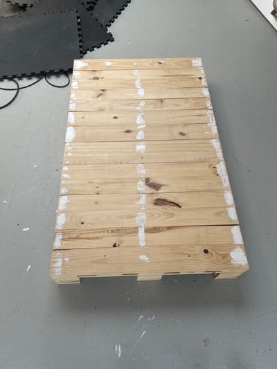 The image shows a wooden pallet with multiple planks. The pallet is lying on a gray concrete floor. Some of the planks have white marks on them, possibly paint or filler. To the left of the pallet are several black interlocking foam mats, and a black cord is partially visible. The scene appears to be outdoors or in a garage due to the concrete floor and the presence of the foam mats, which are often used for exercise or in workshops. The lighting suggests it is daytime, but there are no clear indicators of the time of day or weather conditions. There is no visible text in the image. The pallet seems to be an object in storage or waiting to be used or disposed of.