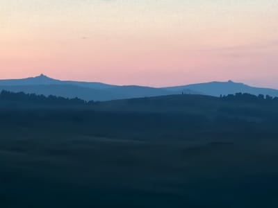 This is a photograph of a landscape at dusk or dawn. The sky is a gradient of pale yellow at the top transitioning to soft pink and then a lighter blue towards the horizon. Silhouetted against the sky are multiple layers of rolling hills. The closest hills are dark blue and appear to be covered in trees. In the distance, lighter blue mountains are visible, with a distinct, pointed peak on the left side of the image. The foreground is a very dark, almost black expanse of land, possibly fields or a valley, with subtle undulating textures suggesting gentle slopes. The overall impression is one of tranquility and vastness. The location is stated as Kuseevo, Russia.