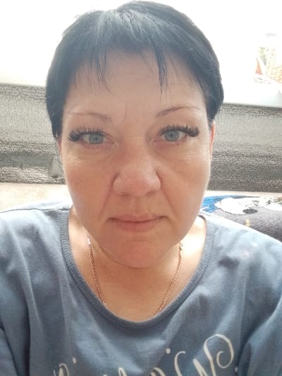The image is a close-up, front-facing selfie of a woman with dark, short hair and blue eyes. She is wearing a light blue t-shirt and a thin gold chain necklace. Her expression is neutral, and her eyes are looking directly at the camera. The background is blurred, but it appears to be an indoor setting with some textured surfaces and what looks like a rug or blanket visible at the bottom. There are no other people or significant objects in the frame. The lighting suggests it may be daytime, possibly near a window due to the brightness. The location is not specified in the image.