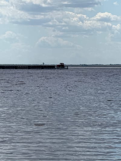 The image depicts a wide, calm body of water under a cloudy sky. In the distance, a long, dark barge is visible, with a small red structure, possibly a cabin or control room, on its right end. A single, slender tree stands on the barge. The barge appears to be moving away from the viewer, or is stationary. The water's surface is textured with horizontal ripples, suggesting a slight breeze. The horizon line is faint, separating the water from a distant shoreline characterized by a low line of trees and a pale sky filled with scattered clouds. The overall impression is one of quiet, expansive openness. No specific landmarks identifiable with the city of Bursol', Russia are present in the image.