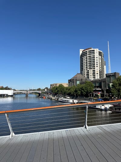 The image shows a cityscape by a river under a clear blue sky. Modern buildings and trees line the riverbank, alongside a walkway with gray planks. Boats are docked along the river, with a bridge visible in the distance. Construction is underway as seen by a crane. The sun is shining brightly, reflecting off the water.