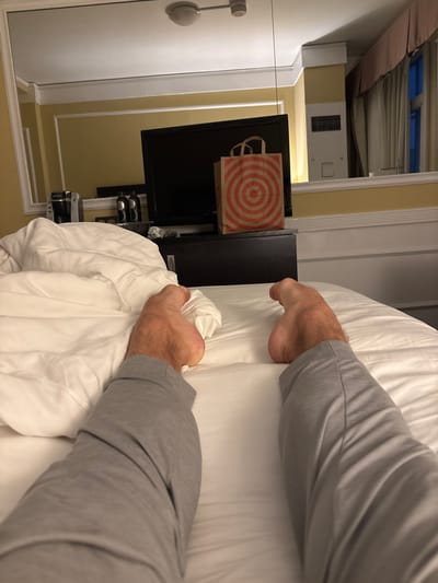 A first-person perspective image shows a person's legs and feet under a white duvet in a hotel room. The person is wearing grey lounge pants. To the left of the person's feet, a white duvet is wrinkled. In the background, a black television is visible, and on top of it is a Target shopping bag. The room appears to be a hotel room with yellow walls and curtains. The image was likely taken in San Francisco, California.