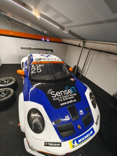 The scene depicts the interior of a pit garage or tent, located in Flatschach, Austria. A white and blue Porsche-style race car occupies the central foreground, angled slightly towards the right. The car features prominent branding including "SenseIT," "GP ELITE," "Sixt," "TAG Heuer," and "MICHELIN," along with the number "25" and the driver's name "A. Levi" on the windshield. A Star of David symbol is visible on the roof. The vehicle appears to have dirt or water marks on its surface, indicating recent use or wet conditions. Its side mirrors are orange, and a large black rear wing is visible. To the left of the car, two Michelin-branded spare wheels are stacked horizontally on the black, interlocking checkered floor matting. The environment consists of a temporary structure with white fabric walls and ceiling, supported by grey metal framework. An orange stripe runs horizontally along the upper wall section. Linear LED light strips illuminate the ceiling. No individuals are present in the image, and no specific actions or interactions are occurring.
