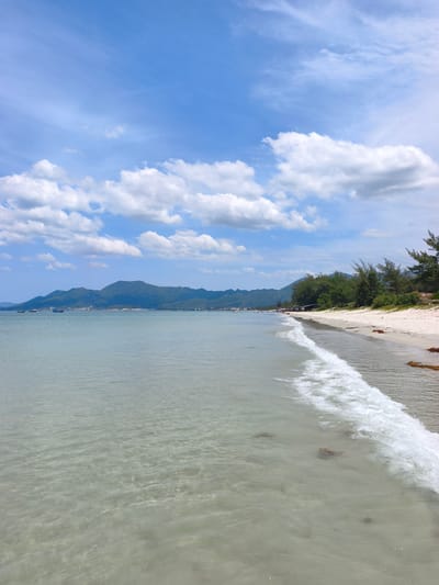 The image captures a scenic view of a tropical beach in the city of Dong Ninh Hoa, Vietnam. The foreground is dominated by clear, shallow water gently lapping at a sandy shore. Small waves with white foam are rolling in, revealing the sandy bottom and a few scattered rocks beneath the surface. The beach itself is a strip of white sand, lined with lush green trees and vegetation. In the distance, a range of rolling, tree-covered mountains forms the backdrop, meeting the horizon under a bright blue sky scattered with fluffy white clouds. A few small boats are visible on the calm sea, and a cluster of people can be seen on the beach in the middle distance, suggesting a popular tourist or local gathering spot. The overall impression is one of a peaceful and idyllic coastal environment.
