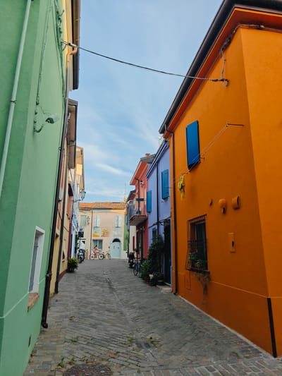The image depicts a narrow, cobblestone street lined with brightly colored buildings in Rimini, Italy. The buildings on either side of the street are painted in vibrant hues of orange, green, and purple, creating a striking visual contrast. The architecture suggests a Mediterranean setting.
The scene appears to be captured in the late afternoon or early evening, with the warm sunlight casting shadows and highlighting the textures of the buildings and the cobblestones. The sky above is a clear blue with wispy white clouds, indicating pleasant weather.
In the distance, two people are visible on bicycles, navigating the street. One person, dressed in pink, is riding a bicycle, and another bicycle is parked nearby. Several motorcycles are also parked along the street. Some potted plants add a touch of greenery to the scene, softening the effect of the bold building colors.
Notable details include the numerous wires and conduits attached to the exteriors of the buildings, suggesting active utility connections. The windows of the buildings feature shutters, some of which are closed. A yellow garment is hanging from a clothesline on the orange building. There are also various wall-mounted fixtures, such as lights and what appear to be electrical boxes.
The overall impression is one of a charming and lively European street, bathed in gentle light. The vibrant colors evoke a sense of cheerfulness and perhaps a holiday atmosphere.
