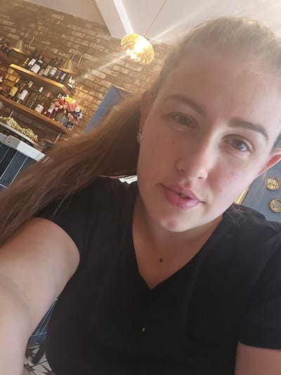 A medium shot selfie shows a woman in a black v-neck t-shirt. She has long brown hair, light-colored eyes, and appears to be wearing light makeup. The background features a brick wall with shelves holding bottles, possibly wine. There is also a decorative light fixture with a metallic, gold-colored shade. The lighting suggests it is daytime. The exact location within the Vale of White Horse, United Kingdom, cannot be determined from the image alone.