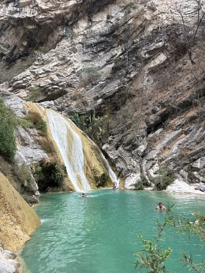 The image depicts a natural outdoor scene in San Miguel Atlapulco, Mexico, dominated by a large waterfall cascading over multi-tiered, yellowish-brown rock formations into a prominent turquoise-colored pool. Steep, stratified rock cliffs, varying in shades of grey, black, and light beige, rise on the right and in the background, exhibiting sparse, dry vegetation and some small green plants. The water in the pool appears clear and calm.
Four individuals are present within the turquoise pool. In the right-center, a male with dark hair is submerged chest-deep, facing towards the left. Near the base of the main waterfall, a person wearing an orange flotation device is floating on their back. Further to the right, at a smaller cascade where water enters the pool, two additional individuals are visible: one person in a red top is wading or sitting directly beneath the descending water, and a second person is partially obscured behind them. Green leafy foliage is present in the immediate lower right foreground, partially obscuring the water's edge.