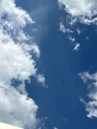 The image shows a clear blue sky with scattered white clouds. A single airplane is visible in the center of the frame, flying at a high altitude. The clouds are puffy and spread throughout the image, creating a dynamic backdrop. The specific location in Manternach, Luxembourg cannot be determined from the image alone.