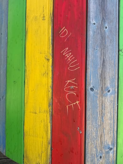 The image shows a close-up of painted wooden planks. The planks are arranged horizontally and are painted in alternating colors: green, grey/blue, red, and yellow. The word "ID NAHUJ KUCE" is carved or scratched into the red plank. The image appears to be taken outdoors and likely in Varkaļi, Latvia.