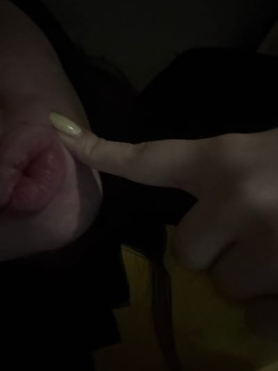 A close-up captures a person's face from a low angle, focusing on their lips and right cheek. A fair-skinned finger with a long, manicured nail presses down on the cheek, creating a dimple. The person has dark hair. The lighting is subdued, casting the scene in shadow.