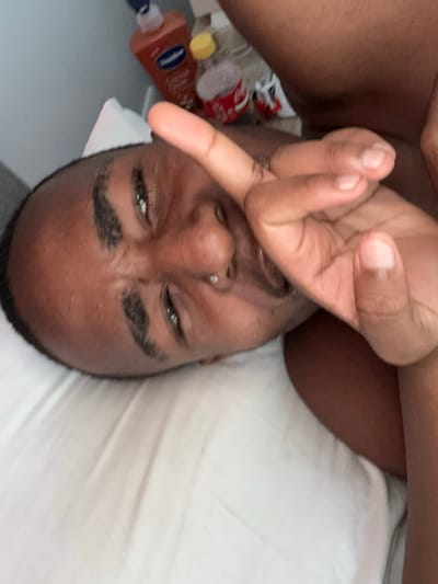 A man with dark skin and short, dark hair is lying on a white bed in what appears to be an indoor setting. He is looking at the camera with a slightly quizzical or annoyed expression. A shiny piercing is visible in his left nostril. His left hand is raised in front of his face with his index finger extended. Next to him on the bed, there is a collection of items including a bottle of lotion and a bottle with a yellow lid. There is no visual information in the image to suggest the location is Pretoria, South Africa.