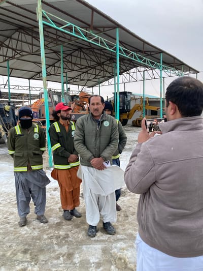 The image captures five men in an open-sided machinery yard in Kan Mehtarzai, Pakistan, beneath a metal shelter with a corrugated roof and light green trusswork. The ground is covered with dirty, melting snow and mud. In the foreground right, a man, seen from behind, wearing a brown jacket over white shalwar, holds a white-cased smartphone horizontally, directing it towards the other four men, presumably recording or photographing them. The central figure, a man with a mustache, wearing a dark olive-green zippered jacket with a green circular emblem and white shalwar, stands facing slightly left, appearing to speak, with his hands clasped, holding a white-cased smartphone. To his immediate left, a man with a red cap and dark hair/beard, wearing a dark olive-green vest with yellow reflective stripes and orange shalwar, stands with clasped hands. Further left, a man in a dark balaclava, dark jacket with reflective stripes, and grey shalwar stands with his hands clasped behind his back. A fifth man is partially visible behind the central figure, wearing a dark beanie and jacket. In the background, under the shelter, several pieces of heavy machinery, including an orange snowplow/loader and a yellow motor grader, are visible. The sky is overcast.