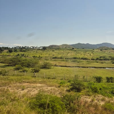 The image depicts a rural landscape in Mkambarani, Tanzania, under a clear blue sky with a few scattered clouds. In the foreground, a dry, grassy area is demarcated by simple fence posts. Beyond this, a gently sloping green field stretches towards a treeline. A narrow, dark body of water or a dry riverbed runs horizontally across the mid-ground, separating this field from another expanse of green vegetation. Further in the distance, rolling hills covered in dense foliage rise to meet the horizon. The vegetation appears lush and green, suggesting a tropical or subtropical environment. The overall impression is one of a tranquil, undeveloped natural setting.