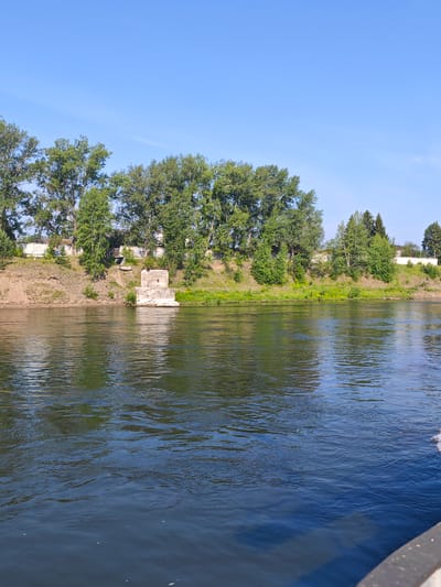 A wide, calm river flows under a clear blue sky on a sunny day. The river's surface reflects the surrounding greenery and the bright sky. On the left bank, a steep grassy slope rises, lined with tall trees. In the middle of the river, a concrete structure, possibly a pier or a remnant of a bridge, stands partially submerged. Beyond the trees on the bank, a white fence is visible, with glimpses of buildings and houses peeking through the foliage. The overall scene is tranquil and depicts a natural landscape with a touch of human infrastructure. The location is identified as Galanino, Russia.