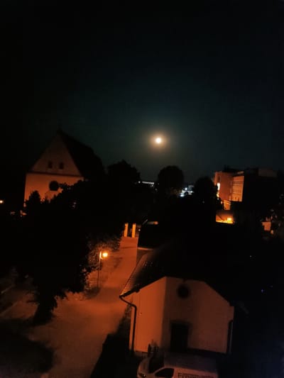 The image is a night time photograph taken from an elevated perspective, likely a balcony or window, overlooking a street in the city of Vyškov, Czechia. The scene is dimly lit by artificial lights and the moon, which is visible in the upper left quadrant of the sky, appearing as a bright, circular orb against a dark, hazy atmosphere.
In the foreground, the roofline of a building is visible, illuminated by an orange glow. To the right of this, a street or pathway is visible, lit by a streetlamp that casts an orange light on the pavement and surrounding trees. The trees are silhouetted against the brighter areas of the scene.
Further back, a prominent triangular-shaped building, possibly a church or historical structure, is visible on the left side of the image. Its facade is illuminated by an orange light, highlighting two small rectangular windows and a circular feature above a dark, recessed entrance. This building is also partially obscured by the shadows of trees.
On the right side of the image, a multi-story building with visible windows emitting light is present. The building appears to be residential or commercial. Other structures and trees are dimly visible in the background, suggesting a moderately urban or suburban environment. There are no visible people or vehicles in motion, creating a still and quiet atmosphere. The overall impression is one of a peaceful night in a Czech city.