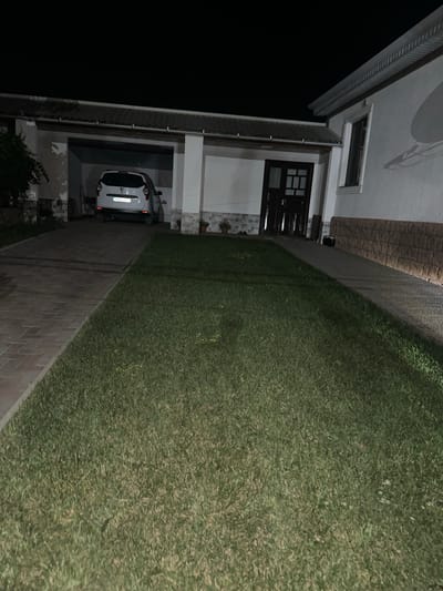 The image depicts the exterior of a house at night in the city of Bolohan, Moldova. A white Dacia Duster is parked under a covered carport, with its rear facing the camera. The house has a light-colored facade, a dark brown door with glass panes, and a window with a dark frame. A paved driveway made of interlocking bricks leads to the carport. Adjacent to the driveway is a well-maintained lawn. The surrounding environment is dark, indicating it is nighttime. The overall scene suggests a residential property.