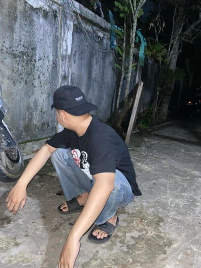A young man wearing a black t-shirt, blue jeans, black sandals, and a black baseball cap is squatting on a concrete ground next to a motorcycle. The man is facing away from the camera, looking to his left. He is in a dimly lit outdoor environment with a concrete wall behind him, covered in patches of moss and stains. Trees and foliage are visible growing along the top of the wall and extending into the darker background. The ground is wet in places, reflecting the ambient light. In the far background, a faint light suggests other vehicles or structures. The scene appears to be in an urban or suburban setting, possibly an alleyway or a less-maintained area in the city of Banjarmasin, Indonesia, given the typical humid climate that would encourage moss growth on walls and the presence of outdoor areas often seen in Indonesian cities.